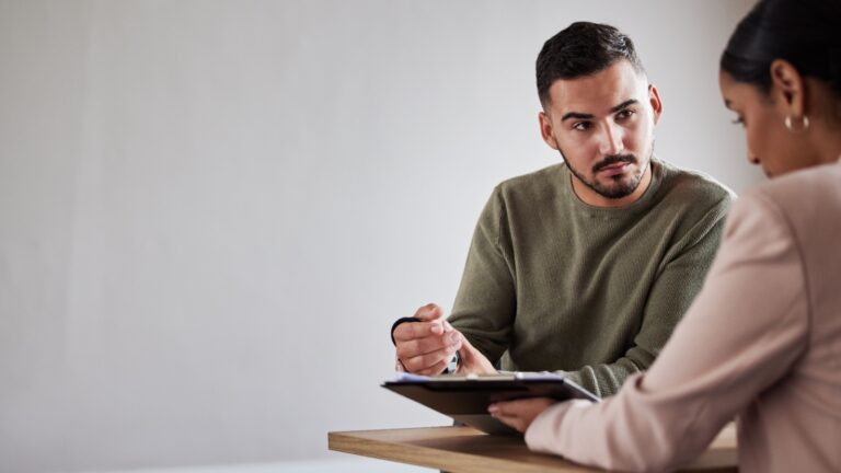 Man holding a clipboard while speaking to a woman sitting across from him.