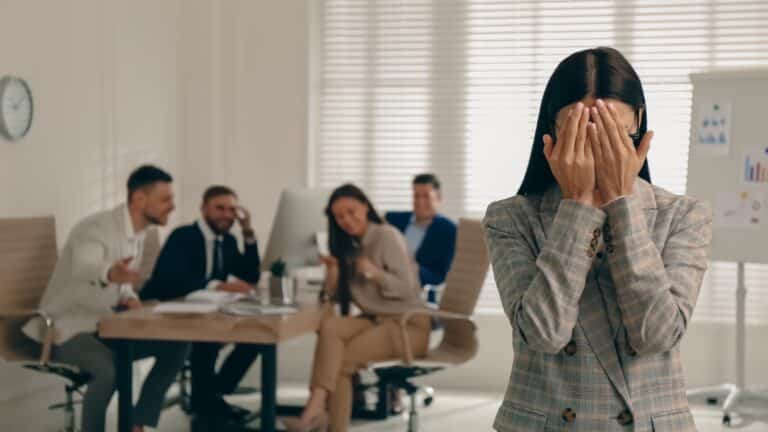 Woman standing alone with hands over her face as a group of employees stare at her. Meant to illustrate workplace harassment.